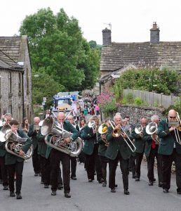 A silver band marching through a village with a trombone player at the front.